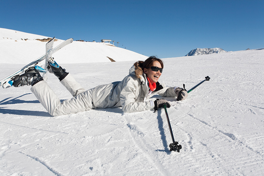 Une skieuse en combinaison est à plat ventre sur la neige, au plat, au soleil. Elle relève les pieds qui sont attachés aux skis. Elle a l'air de s'amuser.