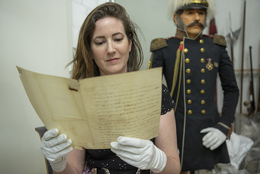 Adelaïde Zeyer tient devant elle, avec des gants blancs, la lettre du général Jomini. Derrière elle, une statue de cire du général en tenue militaire, avec une veste longue à boutons dorés et épaulettes à franges.