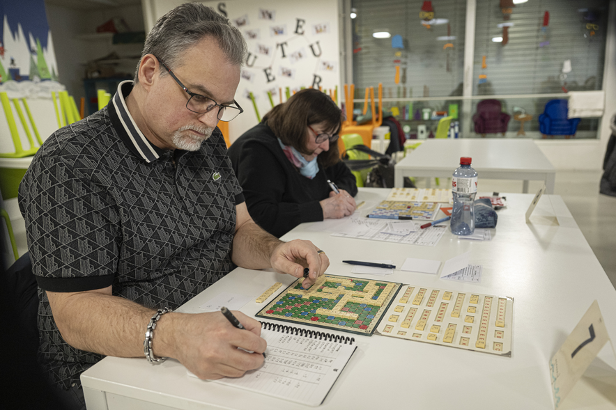 Au premier plan, un homme à lunette est penché sur un plateau de Scrabble. Une femme est assise à côté de lui