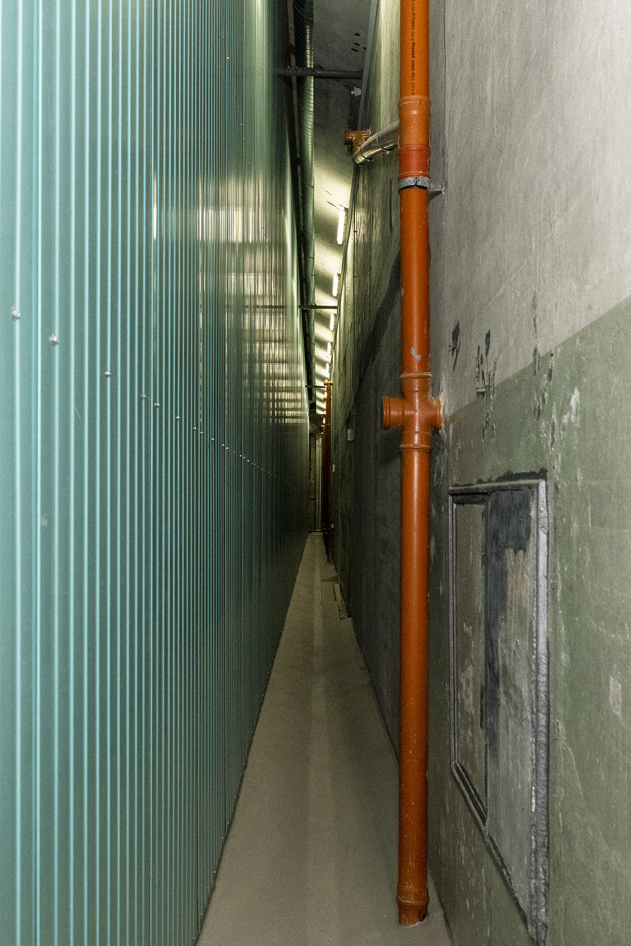 A gauche une longue paroi métallique très haute, à droite le mur de béton à la peinture vieillie, où passe un conduit d'évacuation d'eau. 