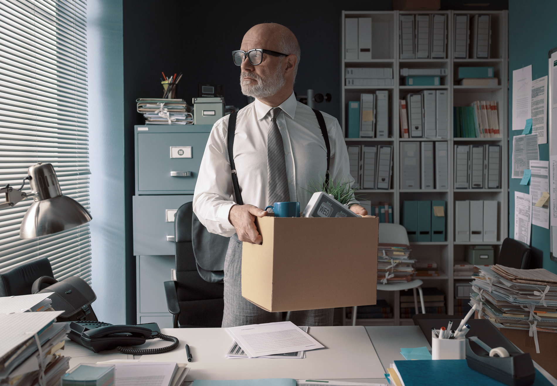 Un homme portant des lunettes, une chemise blanche, une cravate grise et des bretelles, debout derrière un bureau avec des stores à lamelles. Il soulève de son bureau un gros carton duquel on voit dépasser des objets, une tasse et un calendrier.