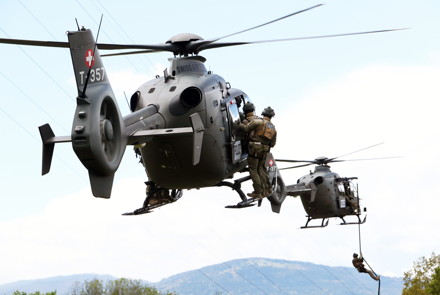 Deux hélicoptères de l'armée suisse dans le ciel. Des agents en uniforme du DARD se tiennent, de dos, sur le marchepied des appareils. Dans l'hélicoptère de devant, un homme est en train de descendre de l'apapreil au moyen d'une corde. Les uniformes sont marqués "DARD" dans le dos de la veste.