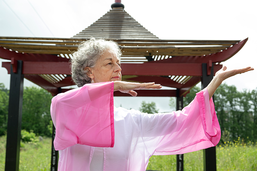 Edith Page, effectuant un mouvement de Qi Gong en tenue traditionnelle rose devant une pergola d'inspiration orientale.