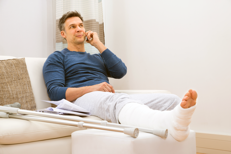 Un homme sur un sofa parle dans son téléphone portable. Il a une jambe dans le plâtre. Une paire de béquilles est posée sur le canapé.