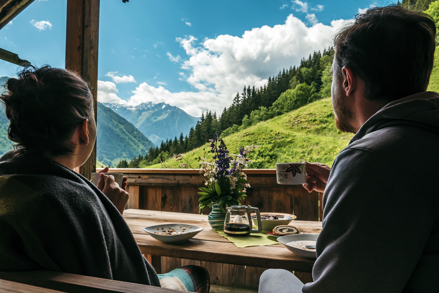 Un couple prend son petit déjeuner assis sur le balcon d'un chalet face à un paysage de montagne et de ciel bleu avec quelques nuages.