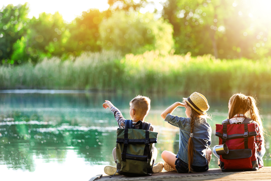 Trois enfants sur un ponton au bord d'un étrang ou d'un lac. On voit des roseaux et des arbres sur l'autre rive.
