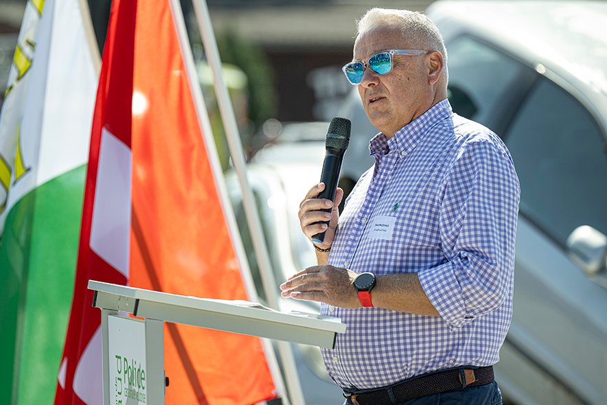 Denis Froidevaux debout à une tribune, en train de parler, avec des drapeaux. Il porte des lunettes de soleil réfléchissantes.