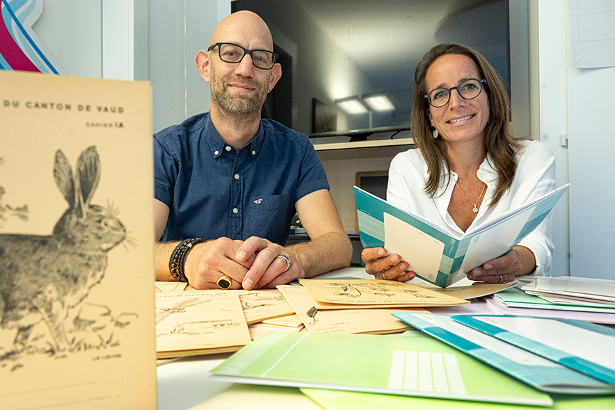 Les deux personnes assises à une table recouverte de cahiers. On voit les anciens cahiers avec leurs couvertures dessinées. Madame Cochard tient un des nouveaux modèles.