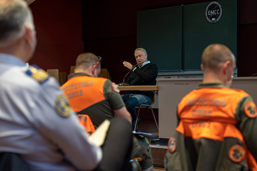 Denis Froidevaux est assis à une table, en train de parler. Au premier plan, des personnes de la protection civile avec des chasubles orange.