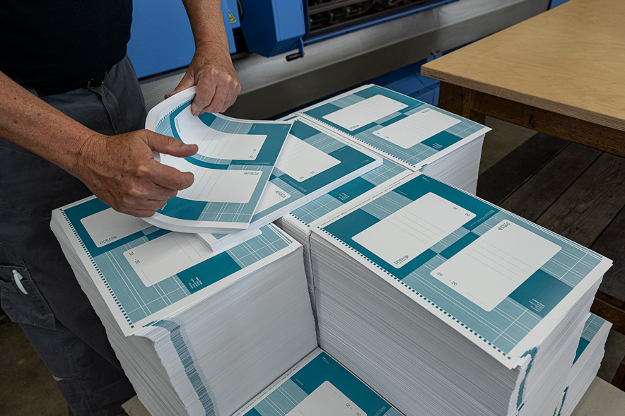 Une personne arrange des piles de feuilles avec au-dessus les couvertures des cahiers, devant une presse d'imprimerie.