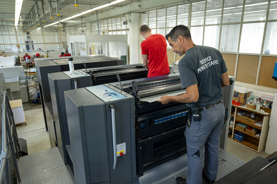 Dans la salle de l'imprimerie des cahiers. Deux personnes s'affairent derrière les machines. L'homme au premier plan porte un t-shirt sur lequel on lit "Service pénitentiaire". Au deuxième plan, un détenu au visage flouté porte un t-shirt rouge.