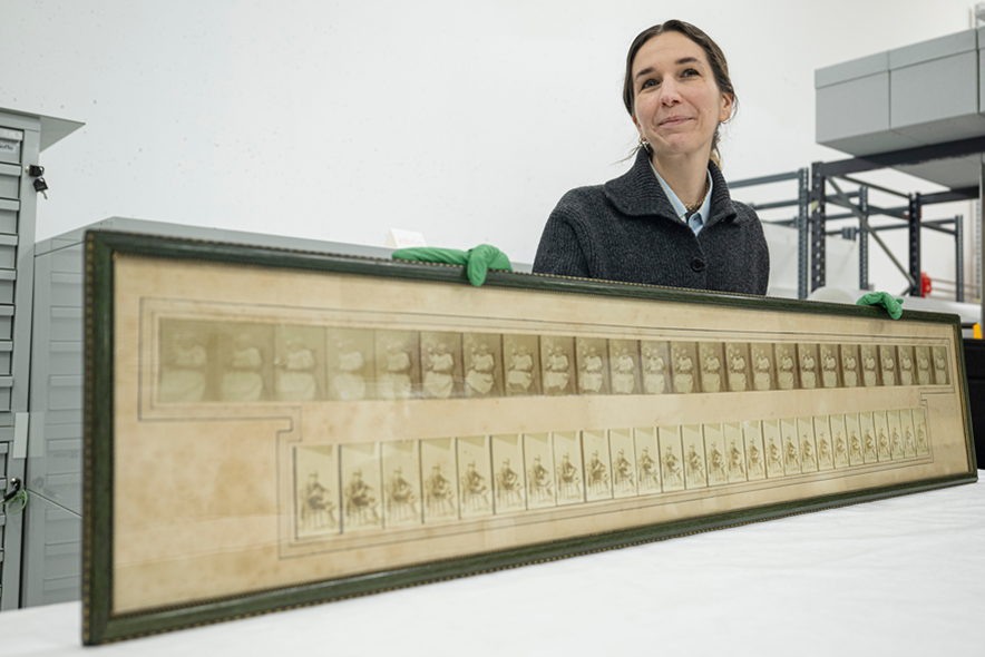 La conservatrice pose derrière un long tableau, une chronophotographie représentant des personnages assis. Elle tient l'objet avec des gants en latex.