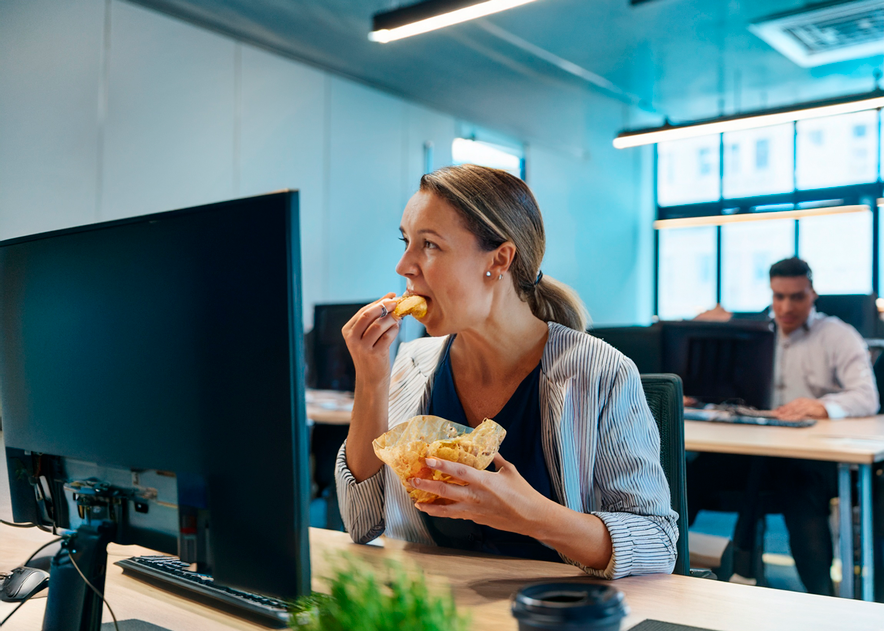 Une femme au bureau, mange devant l'écran de son ordinateur.