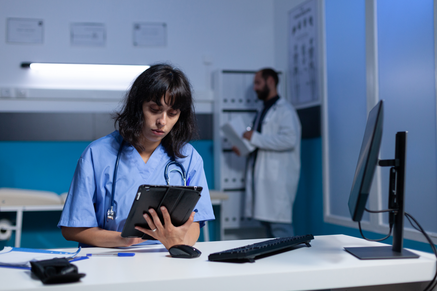 De nuit dans un hôpital: une femme médecin assise à un bureau, travaille sur sa tablette, stéthoscope autour du cou. Au fond, un collègue.