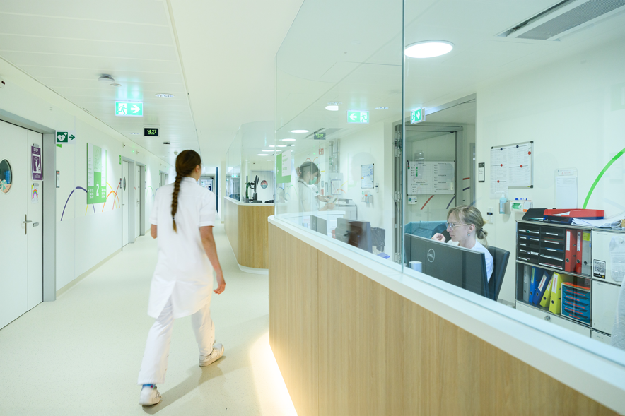 Vue d'un couloir avec un long comptoir de réception à droite. Une femme en blouse blanche marche, de dos. A l'accueil, une personne derrière son écran d'ordinateur
