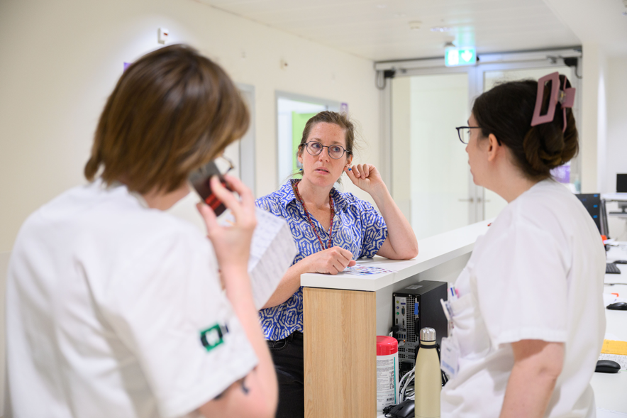 Discussion près d'un desk d'accueil entre deux infirmières ou médecins en blouses blanches et une femme administrative, en chemisier bleu.