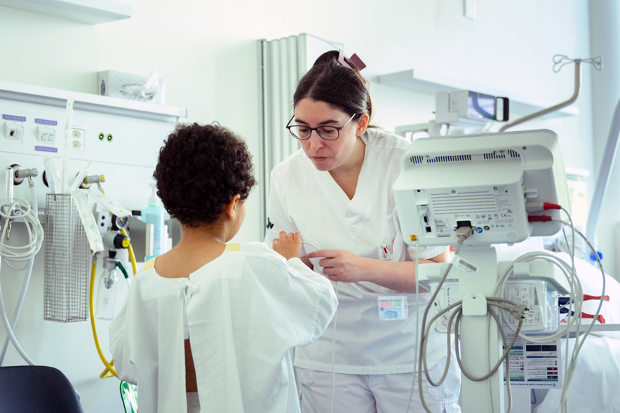 Une médecin inspecte la main d'un jeune patient, de dos, près de son lit.