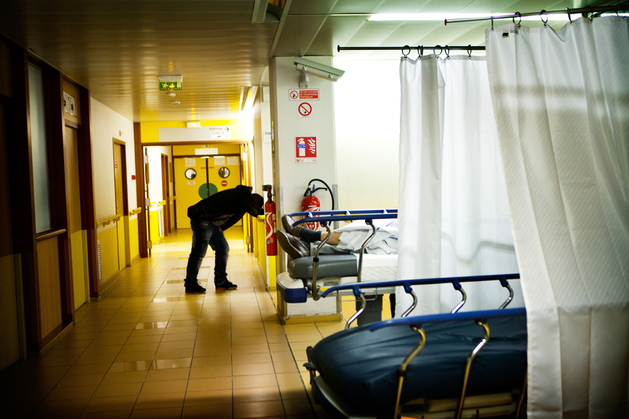 Dans un hôpital la nuit. Au premier plan, des lits d'urgences séparés par des rideaux. Les jambes d'un homme couché dépassent. Au fond, devant ce qui semble un guichet, un homme penché en avant dans une position évoquant la détresse.