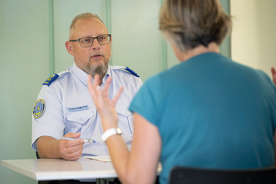 le policier à son bureau fait face à une personne, de dos, qui lève les bras en parlant