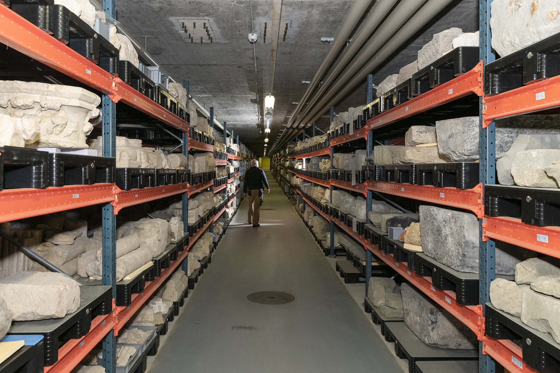 un couloir s'enfonce entre des rayonnages où sont déposés divers objets en pierre. Un homme de dos, arpente le couloir.