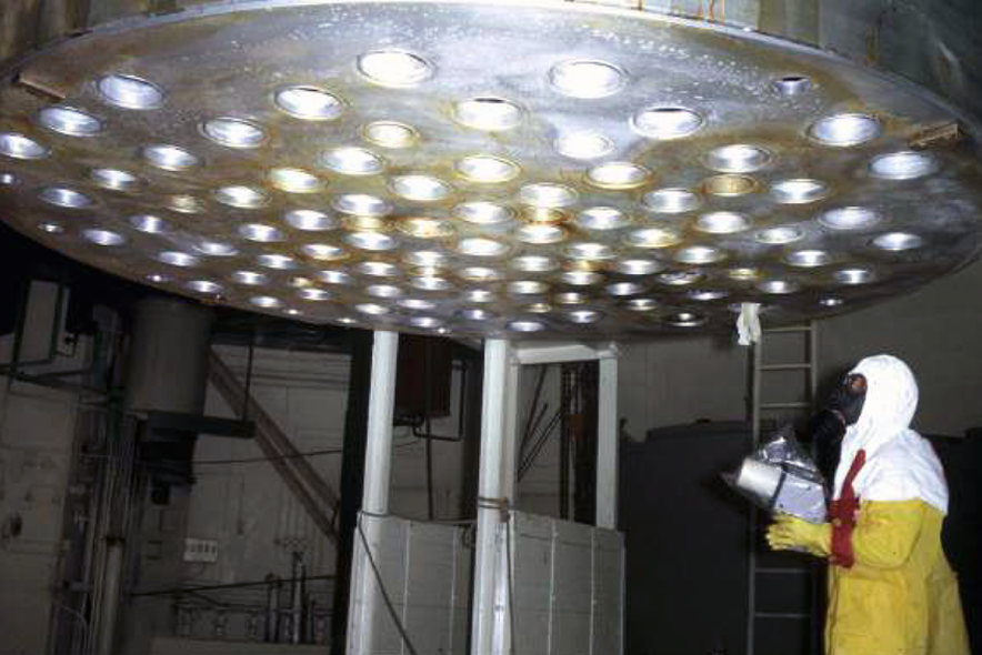 Scientifique en tenue de décontamination portant un masque à gaz et effectuant une inspection sous la cuve de la centrale de Lucens après l'accident de 1969.