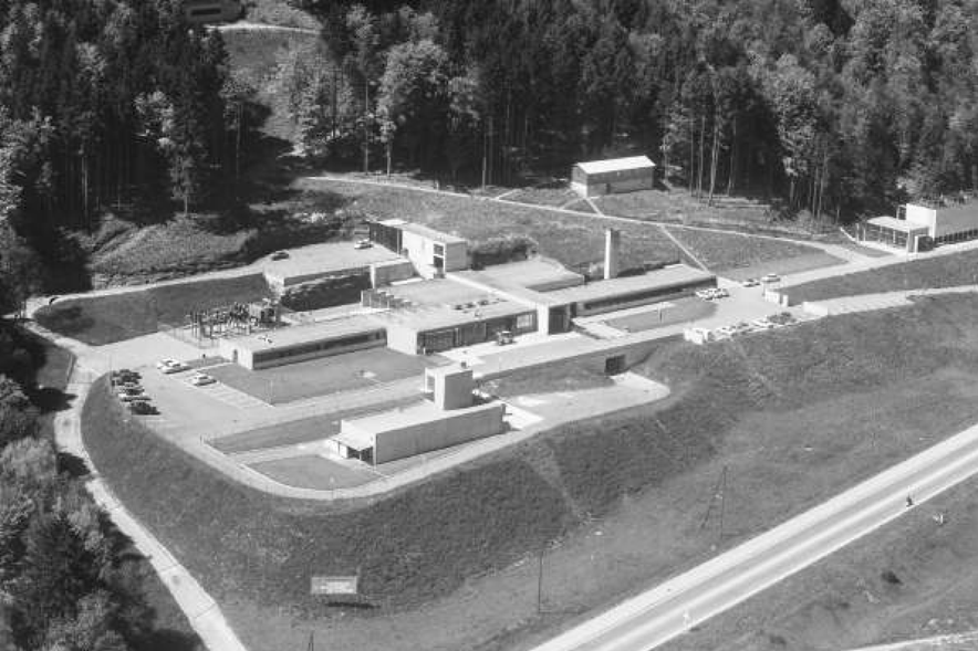 Vue aérienne de la centrale de Lucens : des bâtiments rectangulaires en béton et un parking, le tout sur un talus entouré par la forêt.