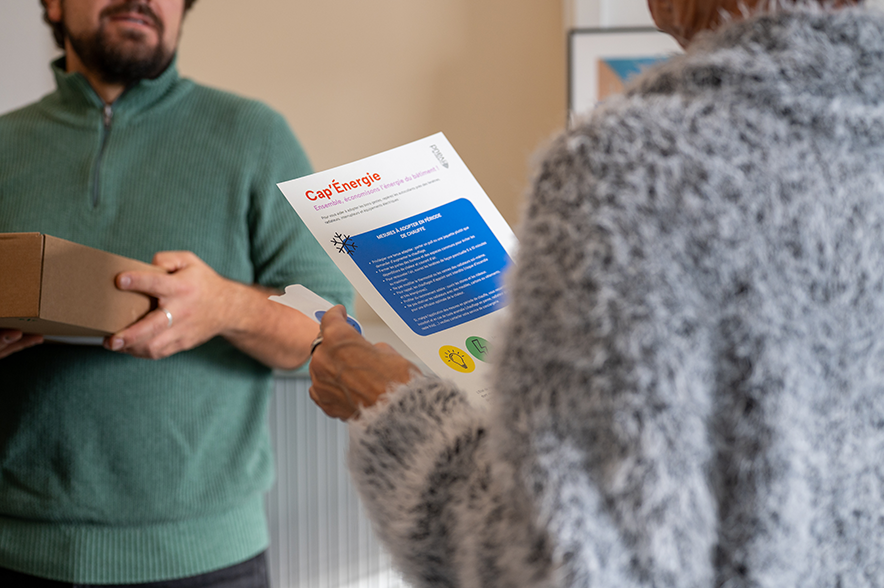 Au premier plan, on voit une personne tenir et lire une affiche avec en face un autre personnage lui donnant des indications.