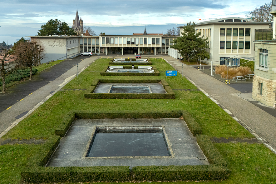 Des fontaines carrées entourés de buis taillés à ras et, au fond, le bâtiment en forme de U, de relativement faible hauteur vu depuis le côté Est.