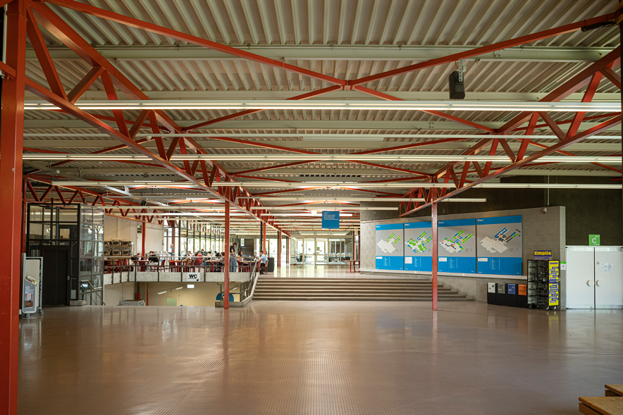 Le hall d'accueil du bâtiment. Un toit métallique supporté par des poutrelles peintes en orange. Au fond, des étudiants attablés. A gauche, un escalier descend. A droite au fond, des panneaux d'affichage.