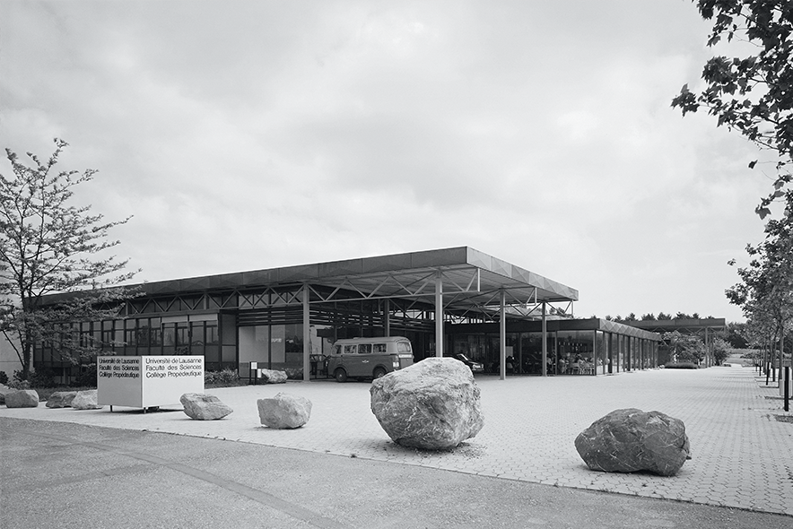 Le parvis du bâtiment avec des pavés de béton et quelques grosses pierres qui empêchent les voitures. Une camionnette VW garée sous un auvent.