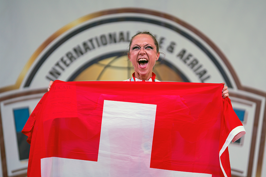 Melody Bacher tient entre ses mains un grand drapeau suisse, dont seule sa tête émerge. Elle fait un grand sourire victorieux. Derrière elle le logo d'une compétition. 
