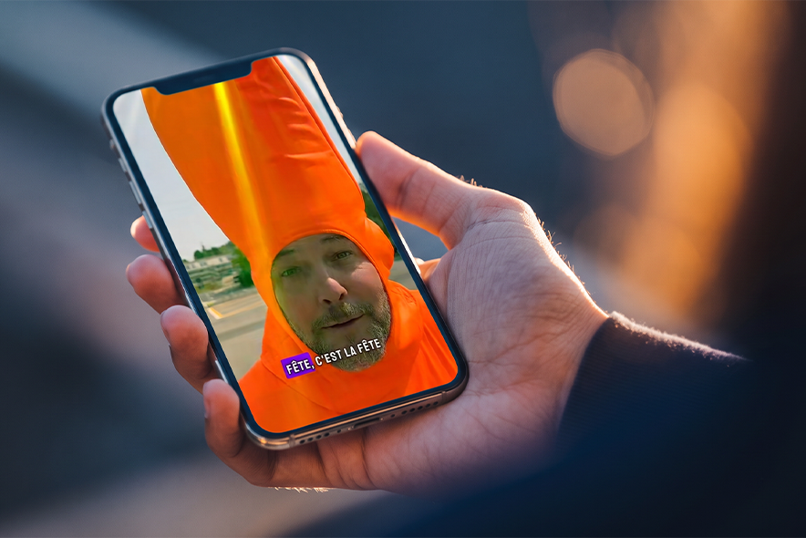 Main tenant un téléphone portable. Sur l'écran, le visage de eCop, coiffé d'une tenue orange vif (il est déguisé en cône de chantier pour une vidéo sur le Tour de France femmes à Lausanne).