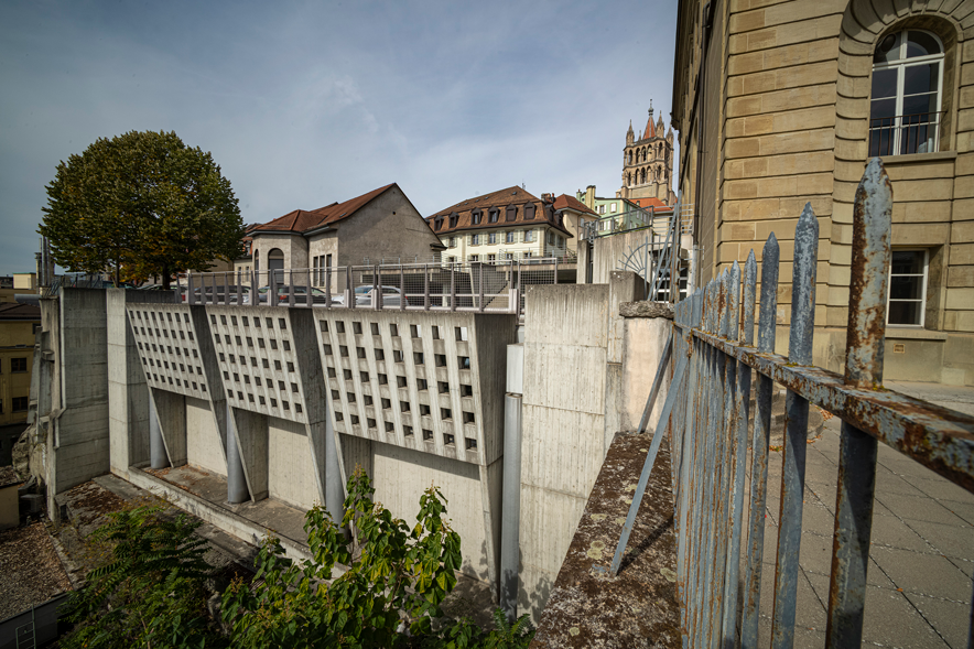 La façade est légèrement oblique, percée de trous carrés réguliers et rythmée par quelques colonnes de béton. Tout à droite de l'image, la clôture et la façade du Gymnase, ancien "Collège scientifique".