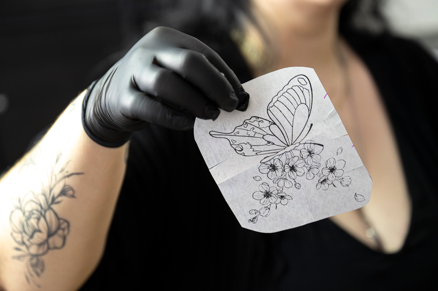 La tatoueuse montre un dessin représentant un papillion qu'elle s'apprête à tatouer