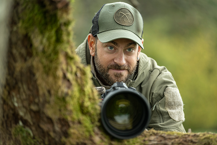 Matthieu Dupertuis en tenue de camouflage, dans la forêt, derrière son appareil photo à l'imposant objectif