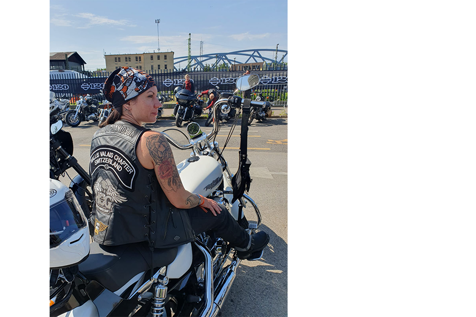 Une femme assise de dos sur une moto. Elle porte un gilet de cuir et un bandana sur la tête.