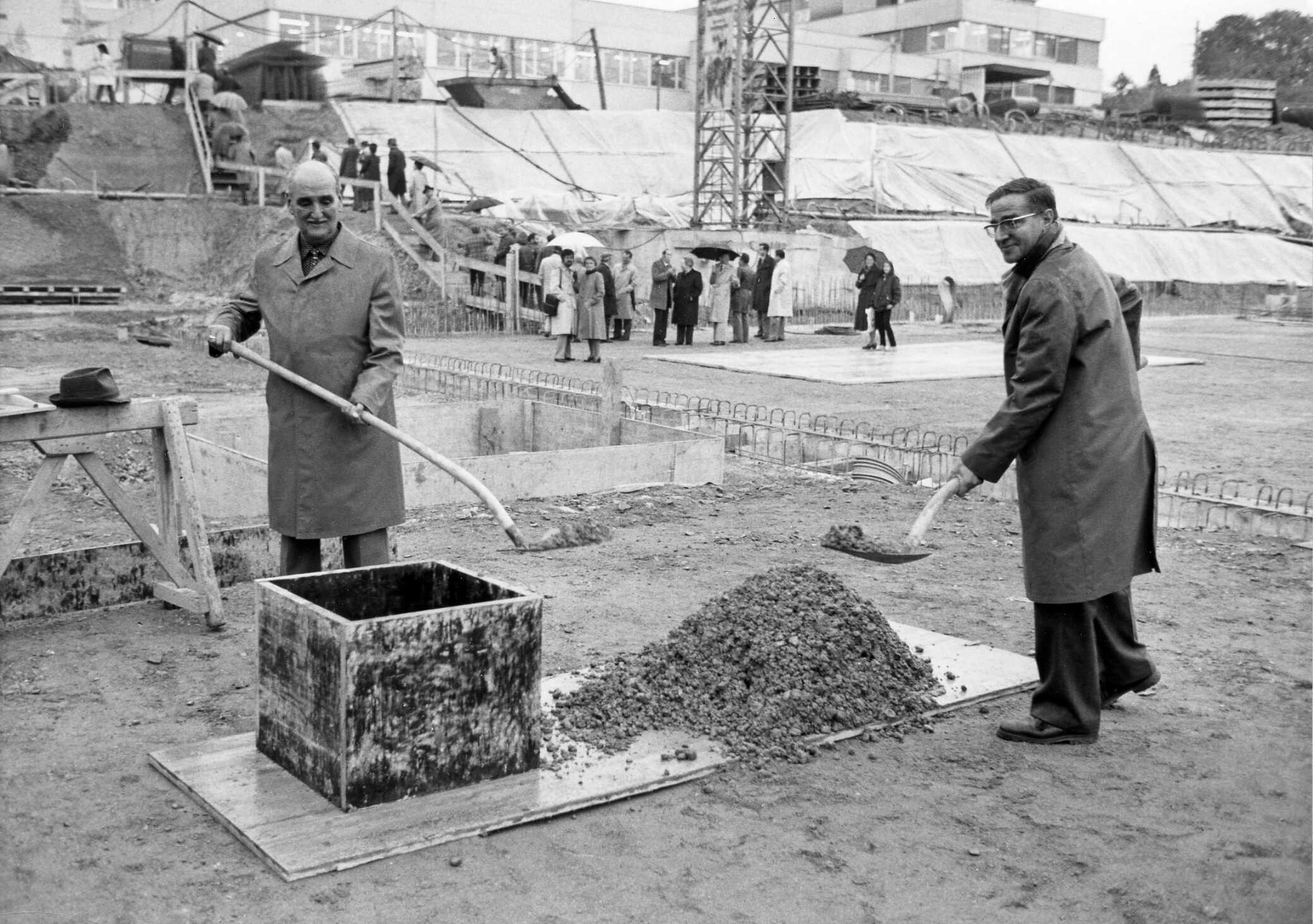 Sous la pluie, deux hommes en manteau sur un chantier portant une pelle. Un groupe de personnes dans le fond