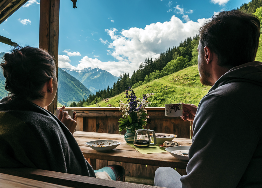 Un couple prend son petit déjeuner assis sur le balcon d'un chalet face à un paysage de montagne et de ciel bleu avec quelques nuages.